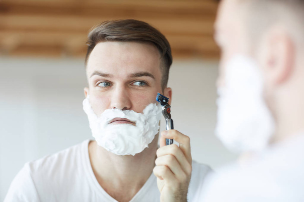 Fej és váll portréja csinos fiatalember borotválkozás reggel nézi a tükörképét tükör, másolási tér - Fotó, kép