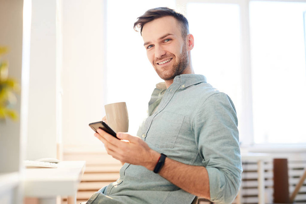 Taillenhoch Porträt eines hübschen jungen Mannes, der es sich am Arbeitsplatz mit einer Tasse Kaffee gemütlich macht und in die Kamera lächelt, Kopierraum - Foto, Bild
