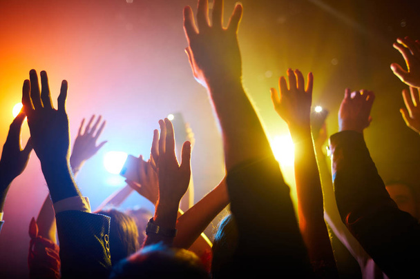 Group of unrecognizable people standing in colorful light and waving hands in air during performance of musician - Photo, Image