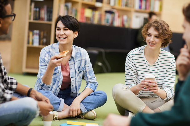 Grupa radosnych podekscytowany przyjaciół studentów siedzi z skrzyżowanymi nogami na podłodze w bibliotece podczas picia kawy i jedzenia pizzy razem - Zdjęcie, obraz