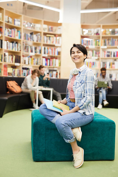 Veselá, sebevědomá studentka s krátkými vlasy na kabelových sluchátkách, které sedí na Pouf v moderní knihovně a drží sešity, a dívá se na kameru - Fotografie, Obrázek