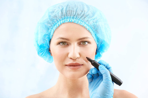 Plastic surgeon applying marking on female face against light background - Foto, afbeelding