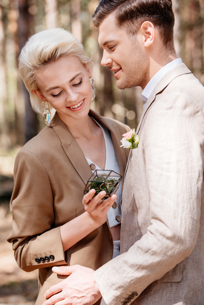 noivo sorridente e noiva segurando caixa com anéis de casamento na floresta
 - Foto, Imagem
