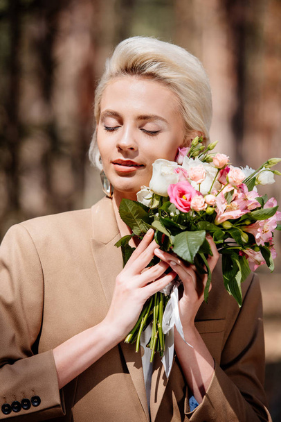 elegant young woman in brown jacket holding bouquet with closed eyes in forest - Photo, Image