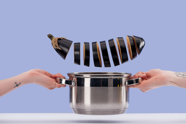 Cropped view of women holding saucepan with freshly sliced aubergine over white surface isolated on blue  - Fotó, kép