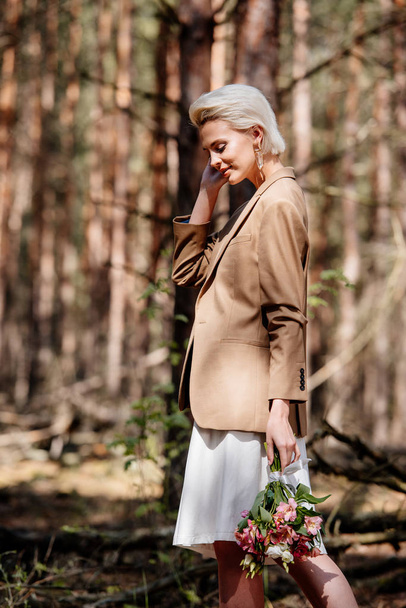 glimlachende blonde vrouw in jas Holding boeket in bos - Foto, afbeelding