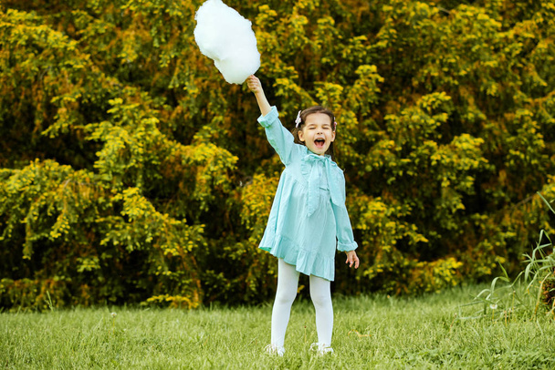 Full body image of happy child eating white cotton candy in the city park. Beautiful little girl wearing light blue dress, having fun outside and posing on the green grass in the holiday. - Photo, Image