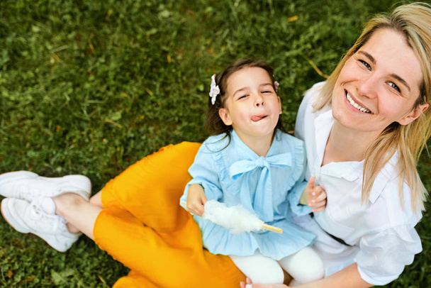 Счастливая мать улыбается и играет со своим ребенком, наслаждаясь время вместе на открытом воздухе. Радостная маленькая девочка ест сладкую вату со своей мамой, сидя на зеленой траве в городском парке. День матери
. - Фото, изображение