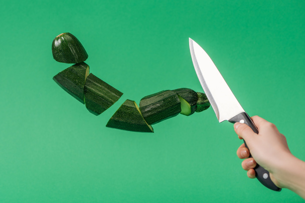 Περικομμένη θέα της γυναίκας που κρατά το μαχαίρι με φέτες φρέσκα πράσινα κολοκυθάκια απομονωμένα σε πράσινο - Φωτογραφία, εικόνα