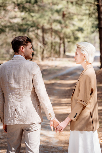 stylish smiling couple holding hands and looking at each other in forest - Photo, Image