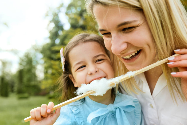 Closeup portrait of happy little girl having fun and eating cotton candy with her mother in the park. Young woman laughing and playing with her joyful daughter, enjoying the time together. Mothers day - Foto, imagen
