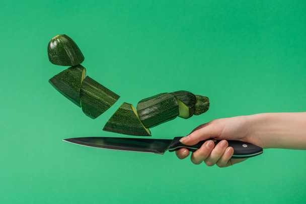Gedeeltelijke weergave van vrouw met mes met gehakte organische courgette geïsoleerd op groen - Foto, afbeelding