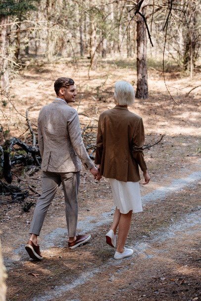 full length view of stylish couple holding hands and walking in forest - Fotografie, Obrázek