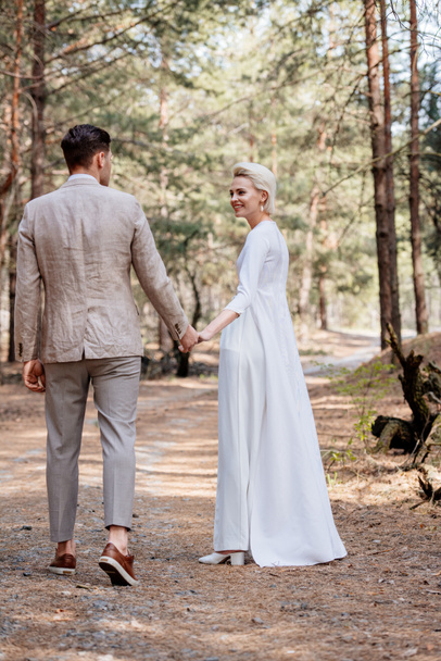full length view of smiling just married couple holding hands and walking in forest - Foto, afbeelding