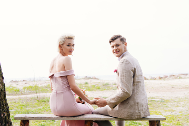 smiling just married couple sitting on bench and holding hands - Photo, Image