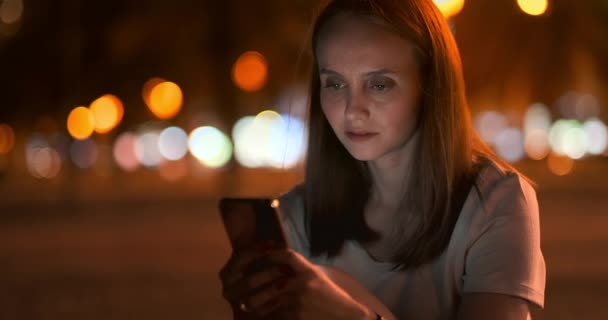 Νεαρό κορίτσι στη νυχτερινή πόλη γράφει μηνύματα κειμένου σε ένα smartphone - Πλάνα, βίντεο