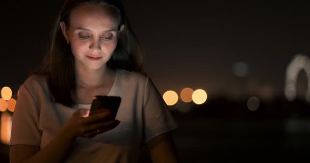 Νεαρό κορίτσι στη νυχτερινή πόλη γράφει μηνύματα κειμένου σε ένα smartphone - Πλάνα, βίντεο