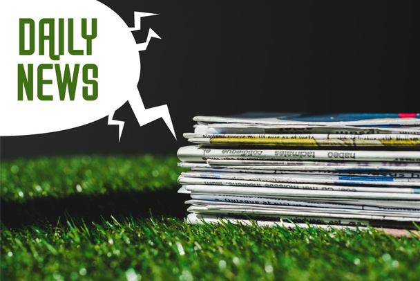 黒で隔離された緑色の毎日のニュースレタリングとスピーチバブルの近くに新鮮な緑の草の上に異なる印刷新聞のスタック - 写真・画像