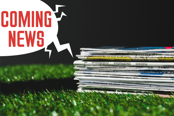 stack of different print newspapers on fresh green grass near speech bubble with red coming news lettering isolated on black - Photo, Image