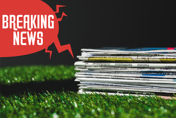 Stapel verschiedener gedruckter Zeitungen auf frischem grünen Gras in der Nähe der roten Sprechblase mit Breaking News Schriftzug isoliert auf schwarz - Foto, Bild