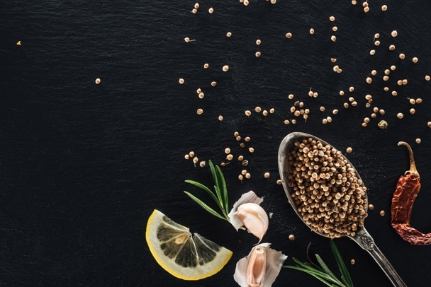 vista superior de semillas de cilantro en cuchara de plata sobre fondo texturizado negro con chile seco, limón, hierbas y ajo
 - Foto, Imagen