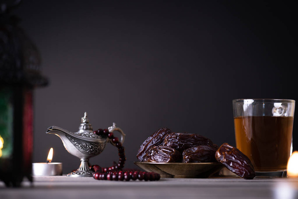Ramadan Essen und Trinken Konzept. Ramadan Laterne mit arabischer Lampe, Holz Rosenkranz, Tee, Datteln Obst und Beleuchtung auf einem Holztisch auf dunklem Hintergrund. - Foto, Bild