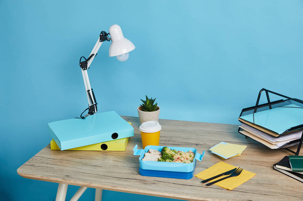 spazio di lavoro con carte sul tavolo e scatola da pranzo con cibo sano su sfondo blu
 - Foto, immagini