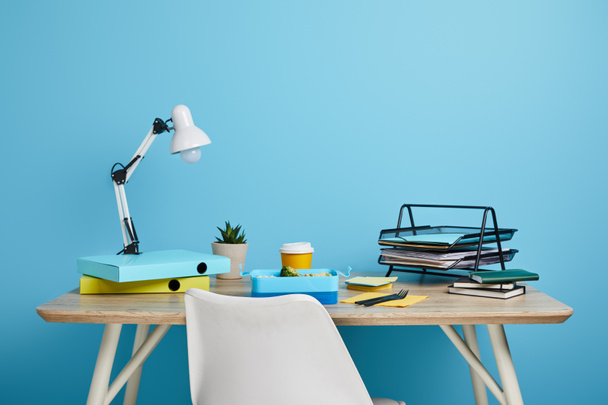 workplace with papers and lunch box with coffee in plastic cup on wooden table on blue background - Fotoğraf, Görsel