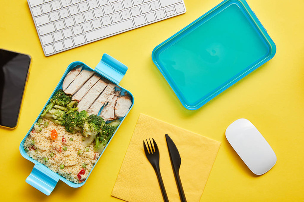 Top uitzicht van smakelijke broccoli met risotto en kip in plastic Lunchbox en computer keyboard op gele achtergrond - Foto, afbeelding