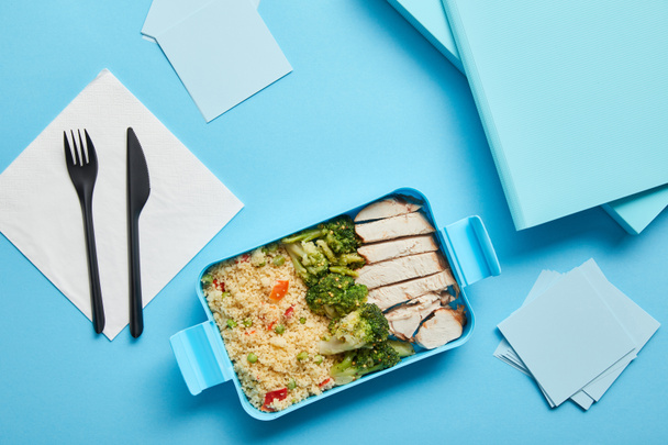 Top view of lunch box with tasty nutritious risotto, chicken and broccoli at workplace on blue background - Photo, Image
