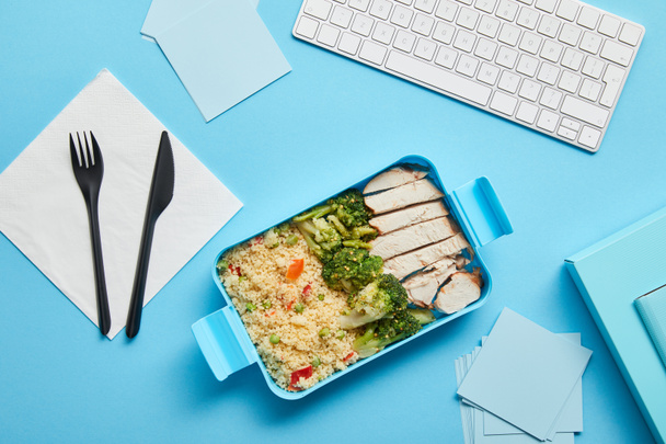 Верхний вид коробки с обедом с вкусным рисом с курицей и брокколи на рабочем месте с клавиатурой компьютера на синем фоне
 - Фото, изображение