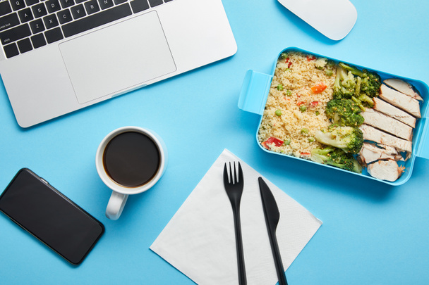 Top view of workplace with digital devices, cup of coffee and lunch box with rice, broccoli and chicken on blue background,illustrative editorial - Photo, Image
