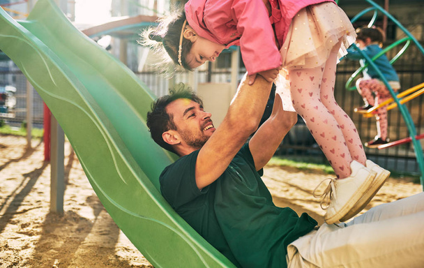 Side view image of cute kid daughter smiling and playing with her father outdoors. Handsome dad and toddler little girl having fun and playing at playground. Father and daughter share love. Fatherhood - Photo, image