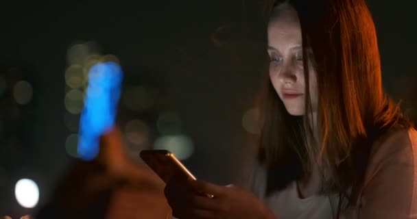 Ένα νεαρό κορίτσι στο παρασκήνιο της νυχτερινής πόλης κοιτάζει στην οθόνη του smartphone - Πλάνα, βίντεο