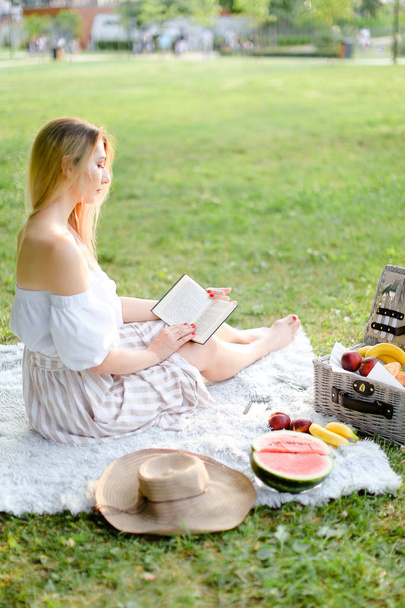 junge blonde Frau liest Buch und sitzt auf Plaid in der Nähe von Früchten im Park. - Foto, Bild