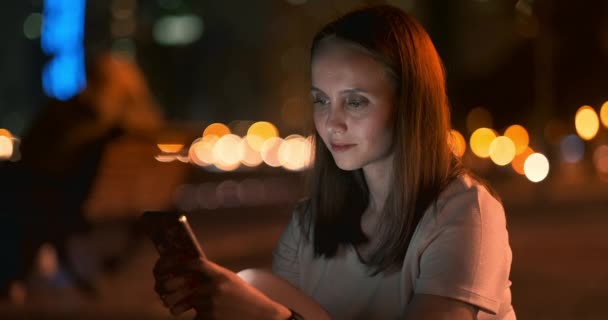 Young girl in the night city writes text messages in a smartphone - Felvétel, videó