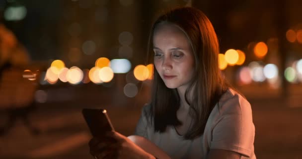Ένα νεαρό κορίτσι στο παρασκήνιο της νυχτερινής πόλης κοιτάζει στην οθόνη του smartphone - Πλάνα, βίντεο