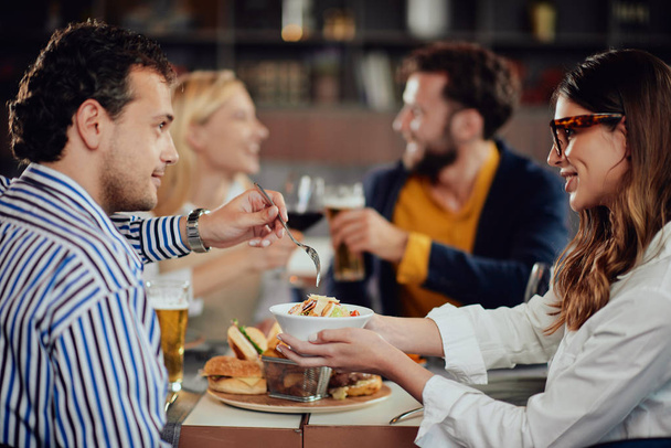 Amigos multiétnicos sentados no restaurante, bebendo álcool, conversando e comendo hambúrgueres para o jantar
. - Foto, Imagem