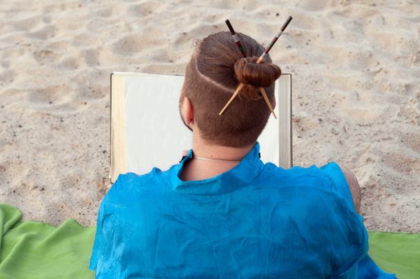 Rückansicht eines bärtigen Mannes mit Dutt und Stöcken auf dem Kopf im blauen Kimono sitzend, Buch am Sandstrand haltend - Foto, Bild
