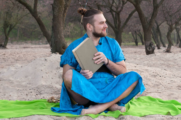 smiling handsome bearded man with make up, bun on head in blue kimono sitting, holding closed book and looking away on sandy beach near tree on rug - Фото, изображение