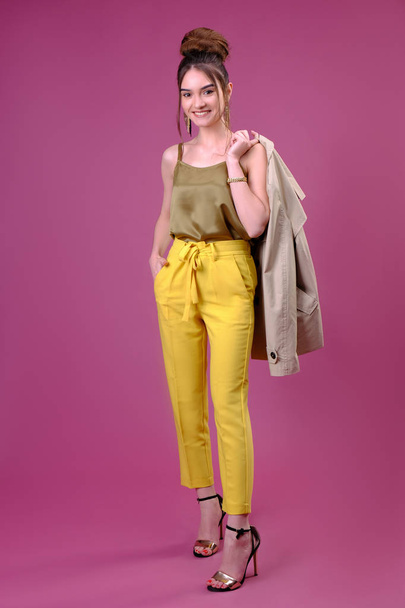 黄色のパンツの若い女性が肩にジャケットを抱えて笑っている。ピンクを背景にした4分の3の長さのスタジオ. - 写真・画像