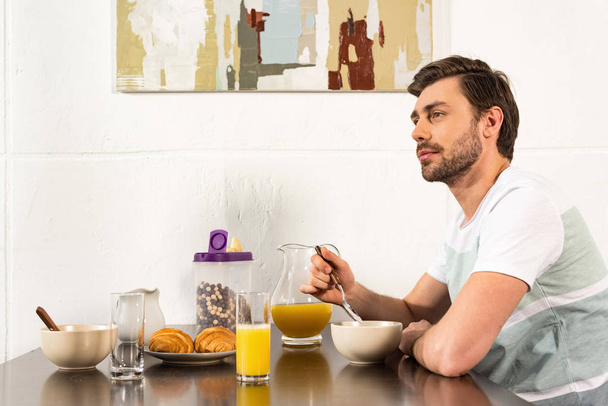bearded man sitting at table during breakfast and dreamy looking away in kitchen - Foto, imagen