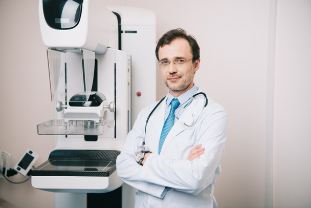 lachende arts staande met gekruiste armen in de buurt van röntgenapparaat en kijken naar camera - Foto, afbeelding