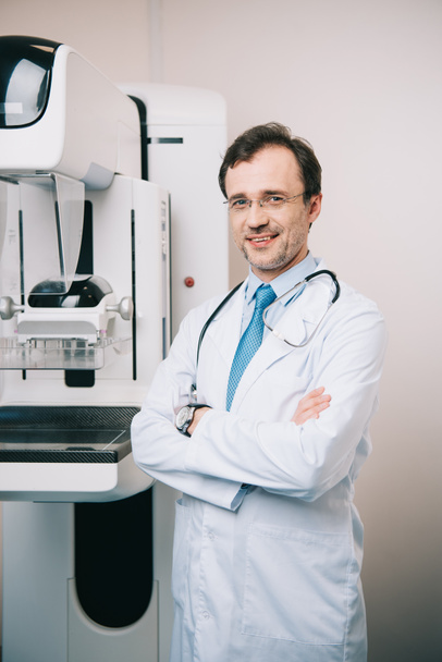Lächelnder Radiologe steht mit verschränkten Armen neben dem Röntgengerät und blickt in die Kamera - Foto, Bild
