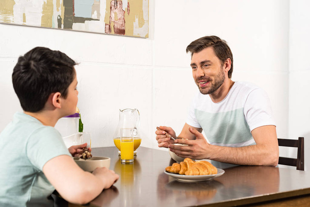 smiling father and son having breakfast and looking at each other in kitchen - Zdjęcie, obraz