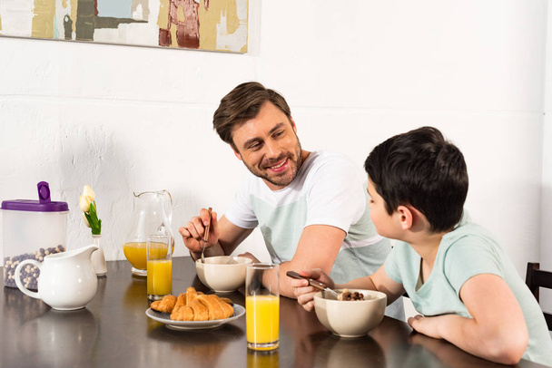 Lächelnder Vater und Sohn frühstücken und schauen einander in der Küche an - Foto, Bild