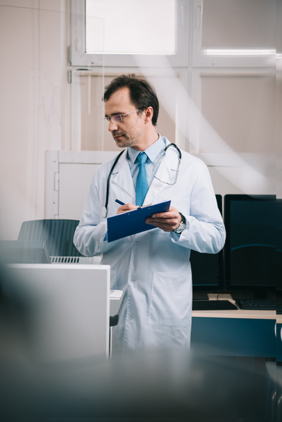 selective focus of handsome doctor in white coat writing on clipboard - Foto, imagen