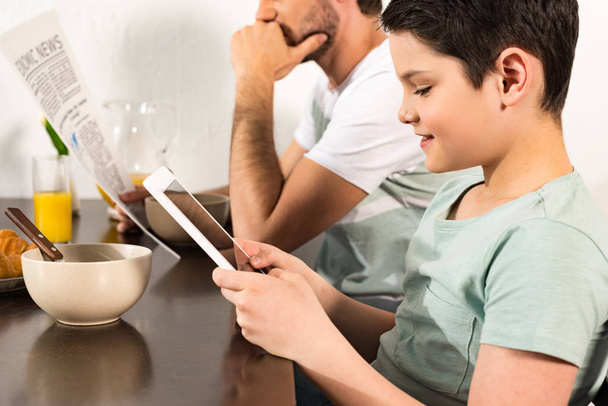 朝食中にデジタルタブレットを使用して新聞を読む父親と息子のトリミングビュー - 写真・画像