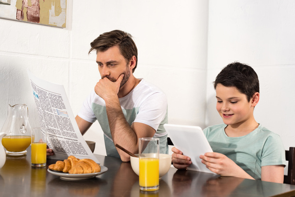 Nachdenklicher Vater liest Zeitung und Sohn nutzt digitales Tablet beim Frühstück - Foto, Bild