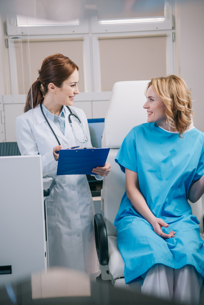 cheerful doctor holding clipboard while standing near patient sitting in armchair - Photo, Image